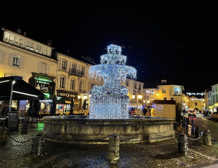 Place de Lattre à Remiremont, de nuit, avec la fontaine illuminée et les commerces